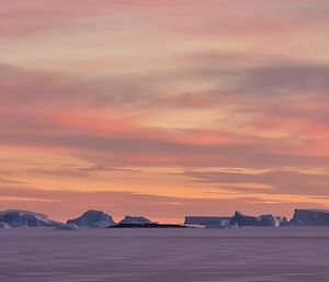 A line of icebergs sit offshore with a dusty pink sky