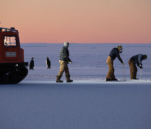 Three men drilling the sea-ice besides hagglunds, with two emperor penguins in background