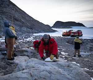 Group in foreground, one smiling at camera with log book open on large rock, others standing and looking at scenery, in distance two hagglunds parked on sea-ice