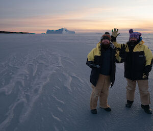 Large expanse of sea-ice with iceberg on the horizen and two men in foreground, dressed in cold weather gear, wearing beanies with pom poms and waving at camera