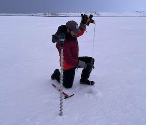 A person kneeling on a wide plain of sea ice. He is inserting a length of stiff measuring tape into a recently-drilled hole in the ice to measure its thickness