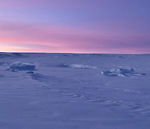An Antarctic dawn, featuring pink-tinted clouds above an irregular snowy landscape. The scene in fact comprises of an ocean bay with icebergs and some low hills in the distance, but they are almost indistinguishable from one another beneath unifying coverings of ice and snow