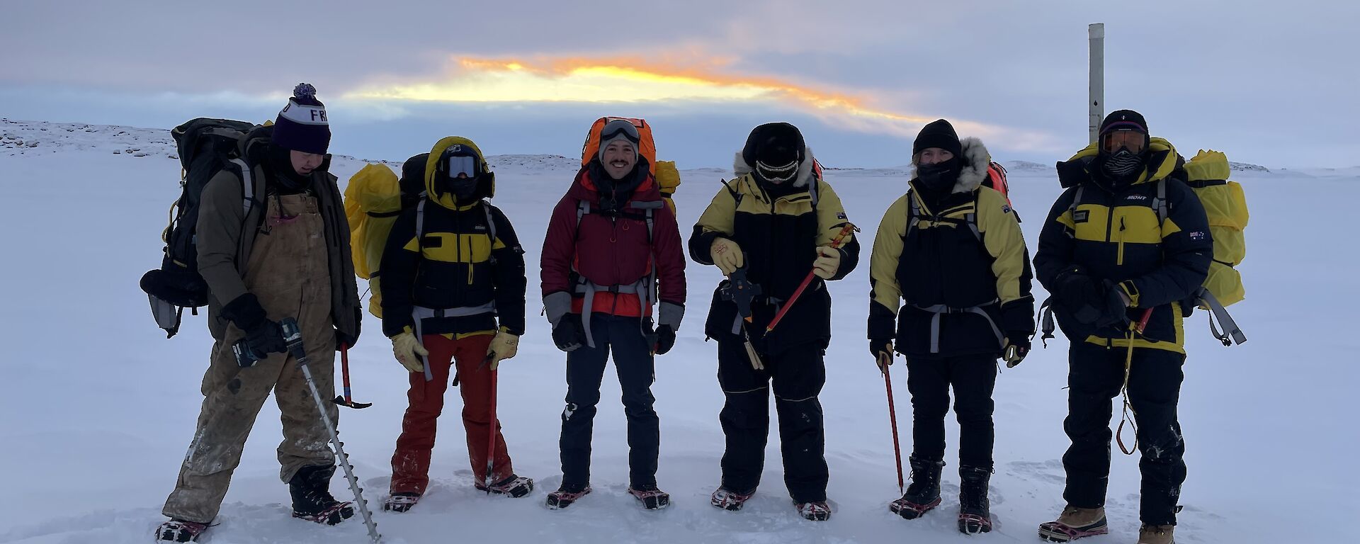 Six people in robust weatherproof clothing including face coverings, thick gloves and padded jackets, standing on a flat, snow-covered stretch of sea ice. They are all wearing large backpacks and carrying ice axes. One holds a powered drill with a metre-long drill flight attached