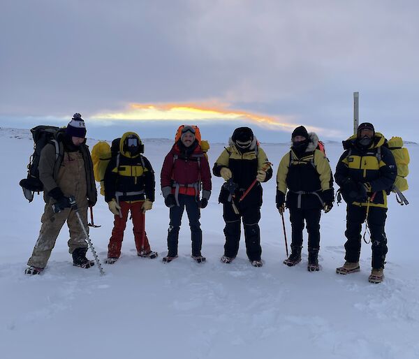 Six people in robust weatherproof clothing including face coverings, thick gloves and padded jackets, standing on a flat, snow-covered stretch of sea ice. They are all wearing large backpacks and carrying ice axes. One holds a powered drill with a metre-long drill flight attached