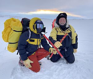 Two people in robust weatherproof clothing including insulated jackets, thick gloves and face coverings, kneeling down on a wide stretch of sea ice. They are each holding up an ice axe, brought together to form an 'X' with the axe handles