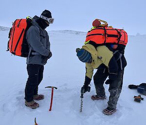 Two people in thick winter clothing standing on sea ice. One is using a powered handheld drill to drill into the sea ice, while the other watches on