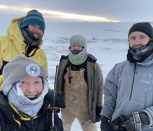 Four people grouped together for a photo, smiling at the camera from behind their neck buffs and balaclavas. They are dressed for very cold weather, also wearing beanies and thick, insulated jackets. A rocky, snow-dusted coastline and sea ice can be seen behind them