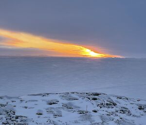 An Antarctic sunset scene. Snow-covered rocks in the foreground slope downwards to a wide expanse of pale, ice-covered ocean. A series of icebergs line the distant horizon. The sunset brings out a streak of fiery colour between the thick clouds overhead and the sea ice below