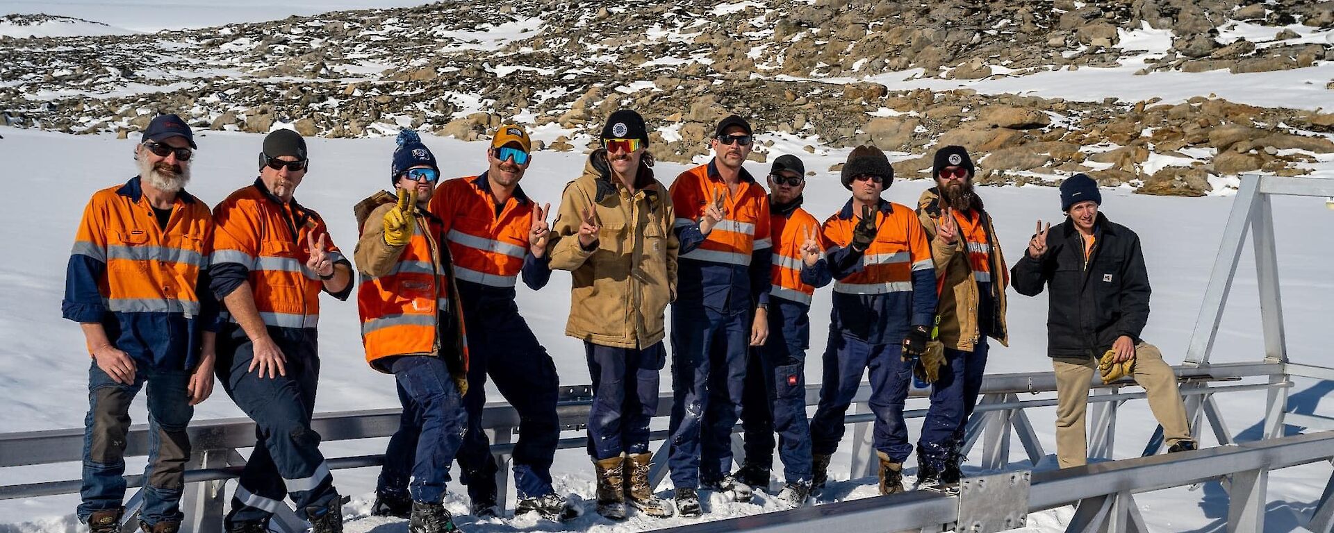 Ten men in high-vis work shirts or jackets, sunglasses and beanies, standing in a row, making 'peace' signs with their fingers. The ground is covered with snow, with a low, rocky hill rising behind them
