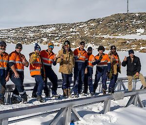 Ten men in high-vis work shirts or jackets, sunglasses and beanies, standing in a row, making 'peace' signs with their fingers. The ground is covered with snow, with a low, rocky hill rising behind them