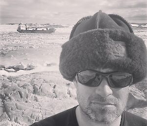 A black-and-white selfie shot of a man in sunglasses and a thick sheepskin hat. Behind him is the ocean, packed with chunks of ice, with a cargo ship moored in the distance