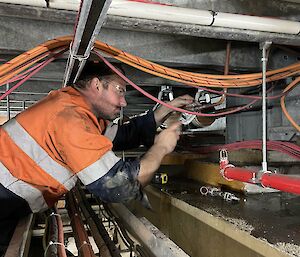 A man in a high-vis work shirt and safety glasses, at work in an under-floor area full of pipes and cables. He is using a wrench to work on a section of piping