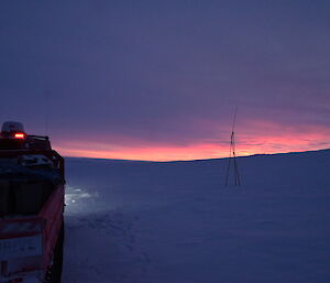 A dawn scene on a snowy plain, taken from just behind a vehicle. The cloudy sky and the ground appear blue-grey against the bright pink and gold light on the horizon