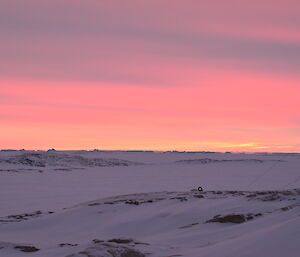 A sunset over a pale, ice-covered ocean with icebergs in the distance. The sea ice appears to be tinted violet under a sky full of high cloud washed with pink and gold sunset colours