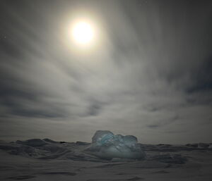 A long-exposure photo of the moon behind a veil of high cloud, above a frozen landscape of irregular ice formations locked into sea ice. The moon appears to be glowing as bright as the sun, though the sky is dark and stars are visible