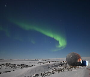 The aurora australis, looking like a band of green light curling at one end, above a snow-covered landscape of low, rocky hills around a frozen ocean bay. A structure shaped like a giant polyhedron stands in the foreground