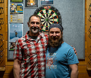 Two men standing in front of a dartboard, arms around each other and smiling for the camera. One wears a plaid shirt, the other wears a bandana and a T-shirt that reads "Bloody Legend 50+ Donations"
