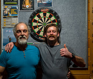 Two men standing in front of a dartboard, arms around each other and smiling for the camera. One of them is giving a 'thumbs-up' gesture