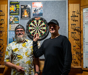 Two men standing in front of a dartboard and smiling for the camera. One wears a shirt with a colourful pattern of prowling tigers, and the other is holding up a dart as if ready to make a throw