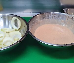 Two metal bowls on chopping board, one with raw onion chopped into rings, one filled with batter