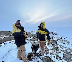 Two men stand over camera on tripod, placed on ridgeline with seaice below and hagglands in distance