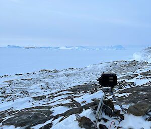 Camera on tripod overlooking rocky outcrop covered in snow but empty of penguins