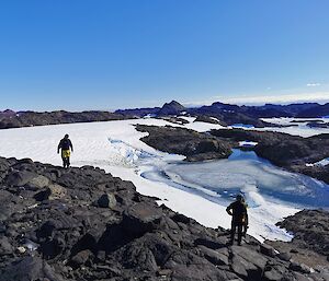 Two people standing on the rocks looking out at snow and a lake