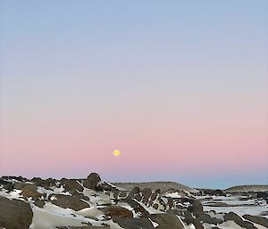 Blue and pink sky with a small moon in the background over snow and rocks