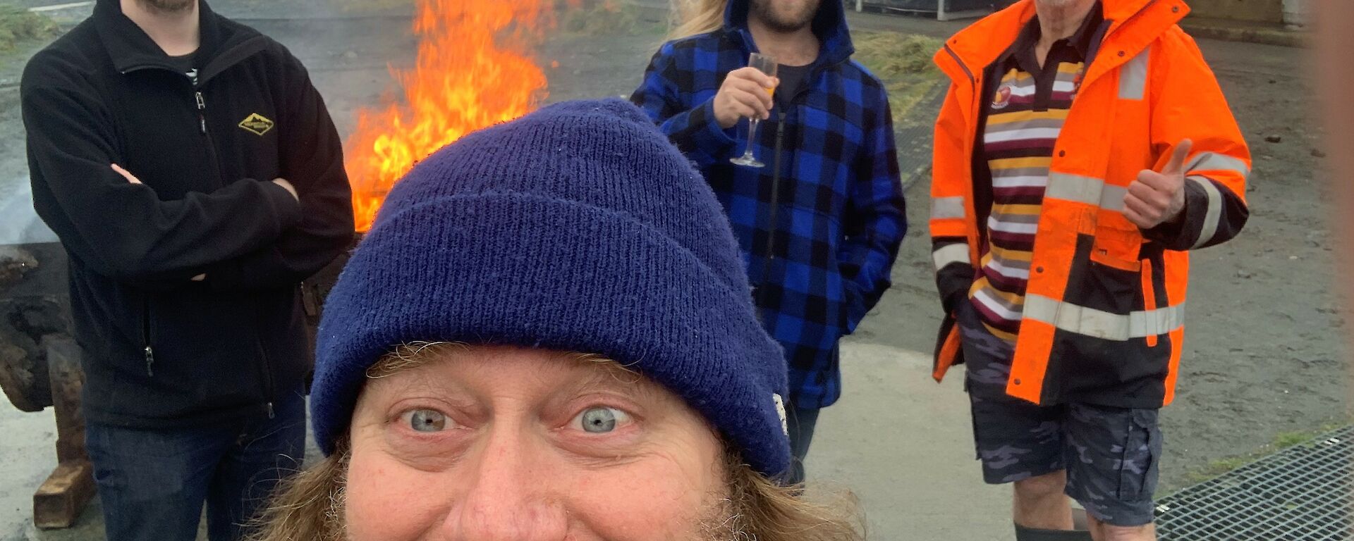 Four men huddled around a fire and smiling. One man is enjoying a glass of sparkling wine.