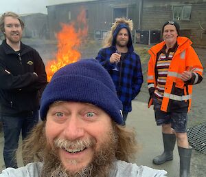 Four men huddled around a fire and smiling. One man is enjoying a glass of sparkling wine.