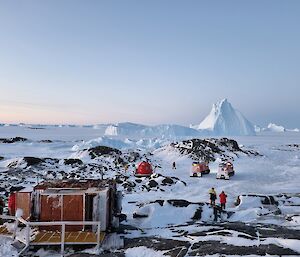 small hut in foreground on rocky snow covered ground, in background peak shaped iceberg