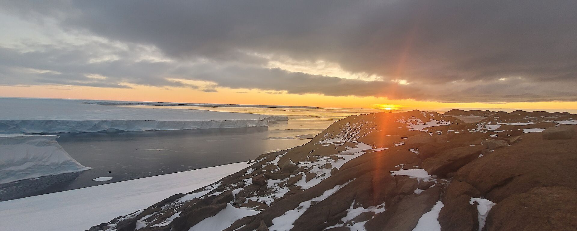 The sun setting over the ocean, beneath low clouds, viewed from some low, rocky hills strewn with snow. Across the water to one side is a glacier, appearing like flat-topped cliffs of ice edging the ocean
