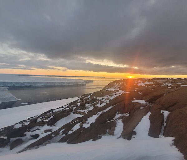 The sun setting over the ocean, beneath low clouds, viewed from some low, rocky hills strewn with snow. Across the water to one side is a glacier, appearing like flat-topped cliffs of ice edging the ocean