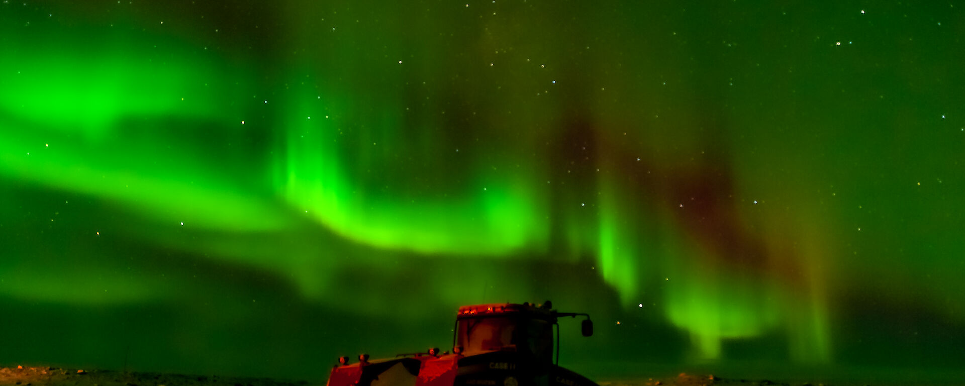The aurora australis filling the night sky with green light varying in intensity, with a duller red stripe just above curving bands of especially bright green. Bright stars can be seen behind the aurora. Below this display is a Quadtrac tractor, parked on a snowy plain