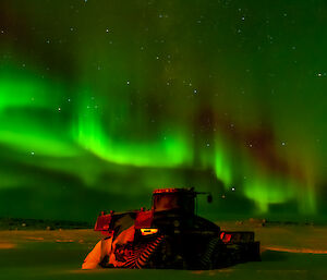 The aurora australis filling the night sky with green light varying in intensity, with a duller red stripe just above curving bands of especially bright green. Bright stars can be seen behind the aurora. Below this display is a Quadtrac tractor, parked on a snowy plain