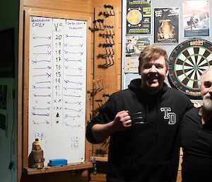 Two men standing with arms about each other, posing for the camera. One man is holding three darts in his closed fist with barbs protruding from between his fingers, to look as if he has claws. Behind the men, a dartboard is mounted on the wall. Behind them to their right is a scoreboard showing scores for teams "Braiden & Steve Casey" and "Dirtbag Diesos Mawson"