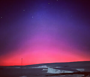 Foreground is east bay with icecliffs and sea ice, and above the horizen the pre-dawn light is bright pink and purple with stars shining above