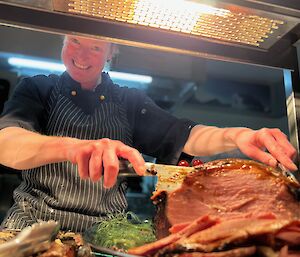 Female chef stands at bea marie smiling at camera while carving a leg ham