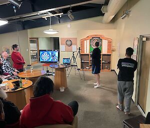 View of the dart board and person throwing the dart from behind with others looking on