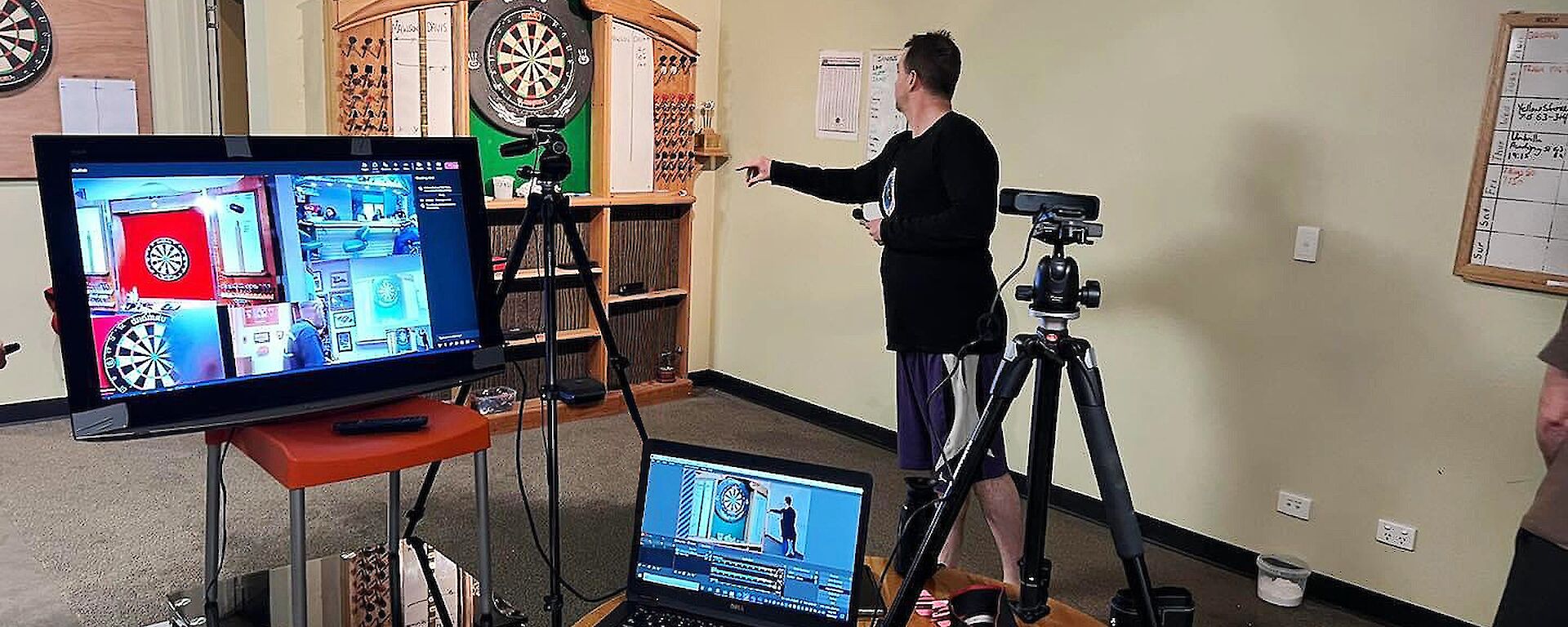 A TV screen, laptop and 2 tripods with cameras with a person throwing darts in the background