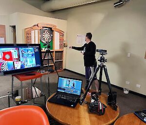 A TV screen, laptop and 2 tripods with cameras with a person throwing darts in the background