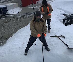 Two men in thick winter work clothing standing on a bank of snow. One is using a long-handled tool to dig a deep hole in the snow. A shovel and a pickaxe are lying nearby