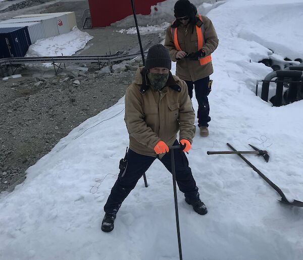 Two men in thick winter work clothing standing on a bank of snow. One is using a long-handled tool to dig a deep hole in the snow. A shovel and a pickaxe are lying nearby