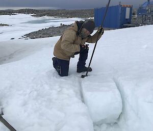 A man in thick winter work clothes and a colourful face buff is kneeling on a bank of snow, holding a long-handled shovel. He is digging out a thick block of snow almost half as long as his own height