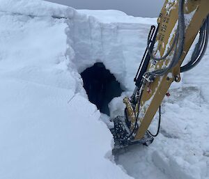 A large bank of snow is being broken up by an excavator. In the process of digging, it has revealed what appears to be a dark cave in the cliff of banked-up snow