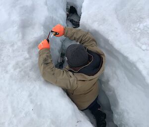 A man wearing thick winter work clothes and holding small electrical tools has descended shoulder-deep into a crevice dug into the snow. He is kneeling in front of an electrical termination box down in the crevice