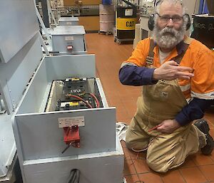 An electrician wearing a work shirt, overalls and ear muffs is kneeling in front of a large battery. He is holding the probes of a multimeter in one hand and making a sign language gesture with the other