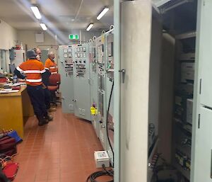 Three men in orange work shirts inspecting meter readings and equipment in a long room lined with electrical distribution boards