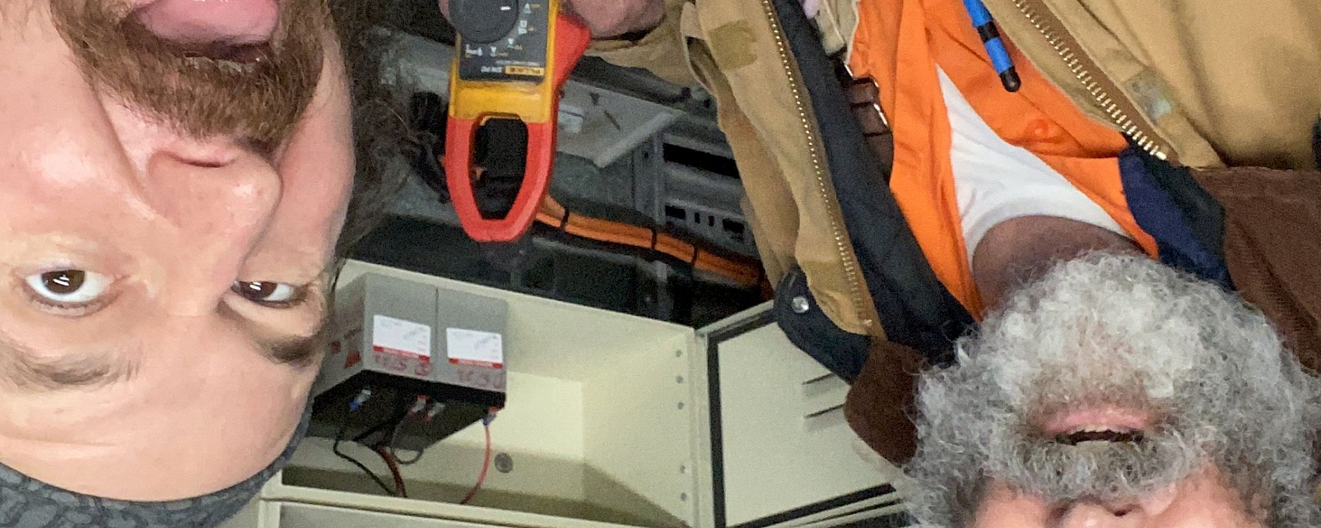 Two electricians standing in front of an electrical alarm panel, one of them holding a multimeter. They are both making comical, open-mouthed faces at the camera