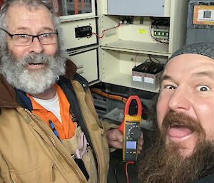 Two electricians standing in front of an electrical alarm panel, one of them holding a multimeter. They are both making comical, open-mouthed faces at the camera